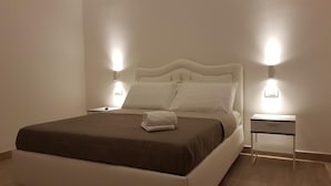 Una cama bien hecha con colcha marrón y almohadas blancas, flanqueada por dos mesitas de noche con lámparas, en una habitación minimalista con paredes de color claro y suelo de madera.