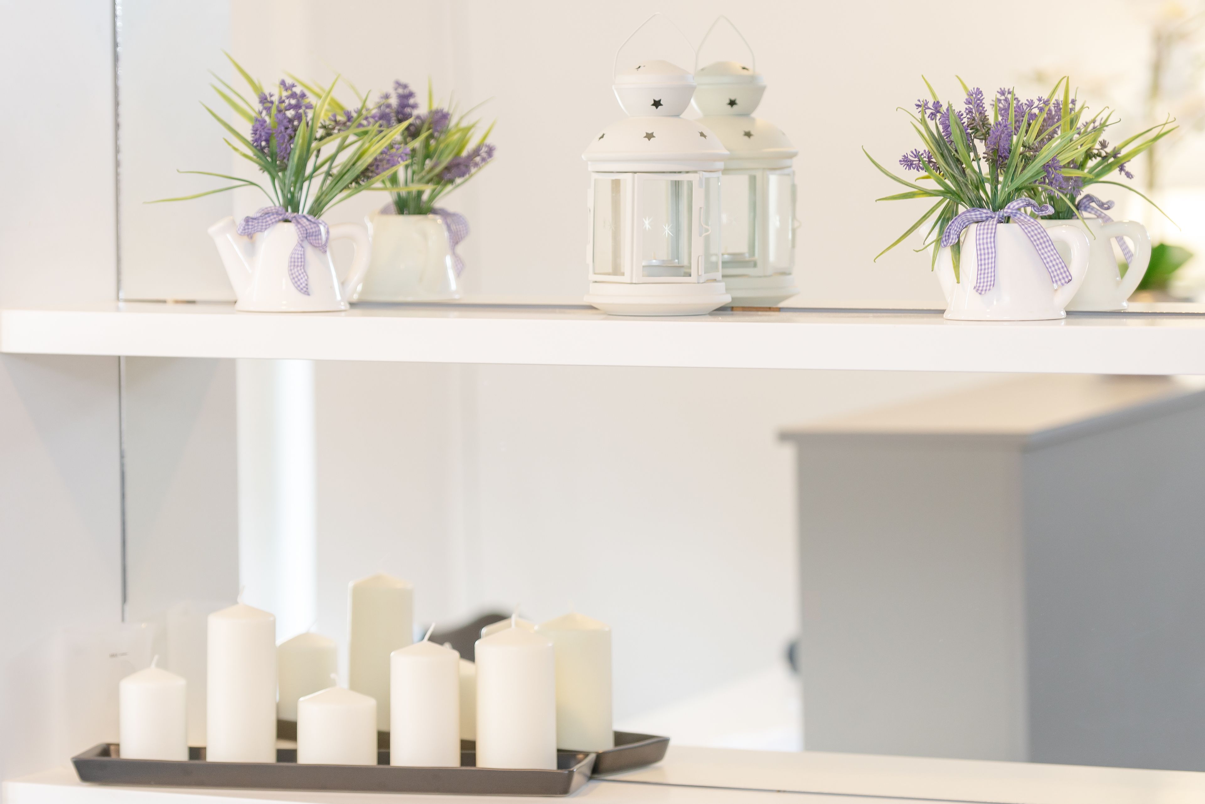 A shelf with white ceramic pots containing purple flowers, white lanterns, and a tray with white candles.