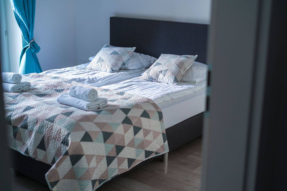 A neatly made bed with a geometric patterned bedspread and pillows, a dark headboard, and a window with curtains.