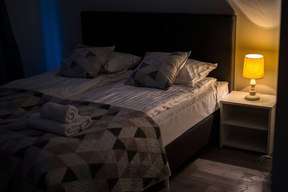 A neatly made bed with a geometric-patterned blanket and pillows, a bedside lamp, and a wooden floor.