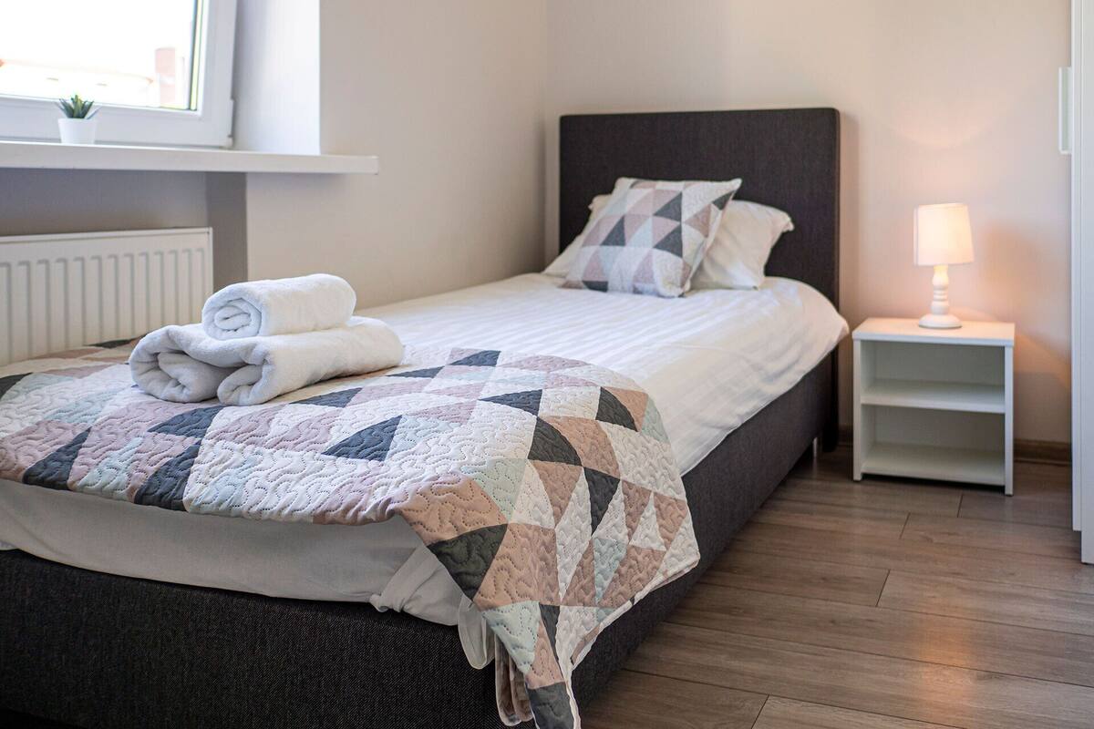 A neatly made bed with a quilt, pillows, and a rolled towel. There is a bedside table with a lamp and a small shelf.