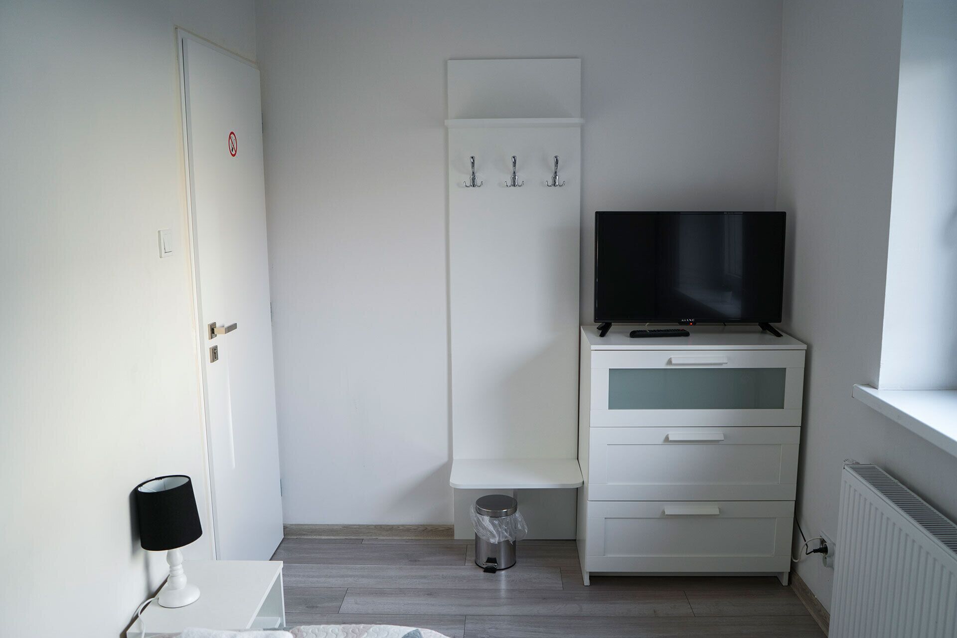A small, clean room with a white wardrobe, a television on top, a small trash bin, and a lamp on a table.