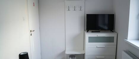 Una habitación pequeña y limpia, con un armario blanco, un televisor encima, un cesto de basura pequeño y una lámpara sobre una mesa.