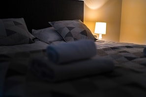 Una cama bien hecha con almohadas estampadas y una toalla enrollada, una lámpara de noche y una pared con una fuente de luz cálida.