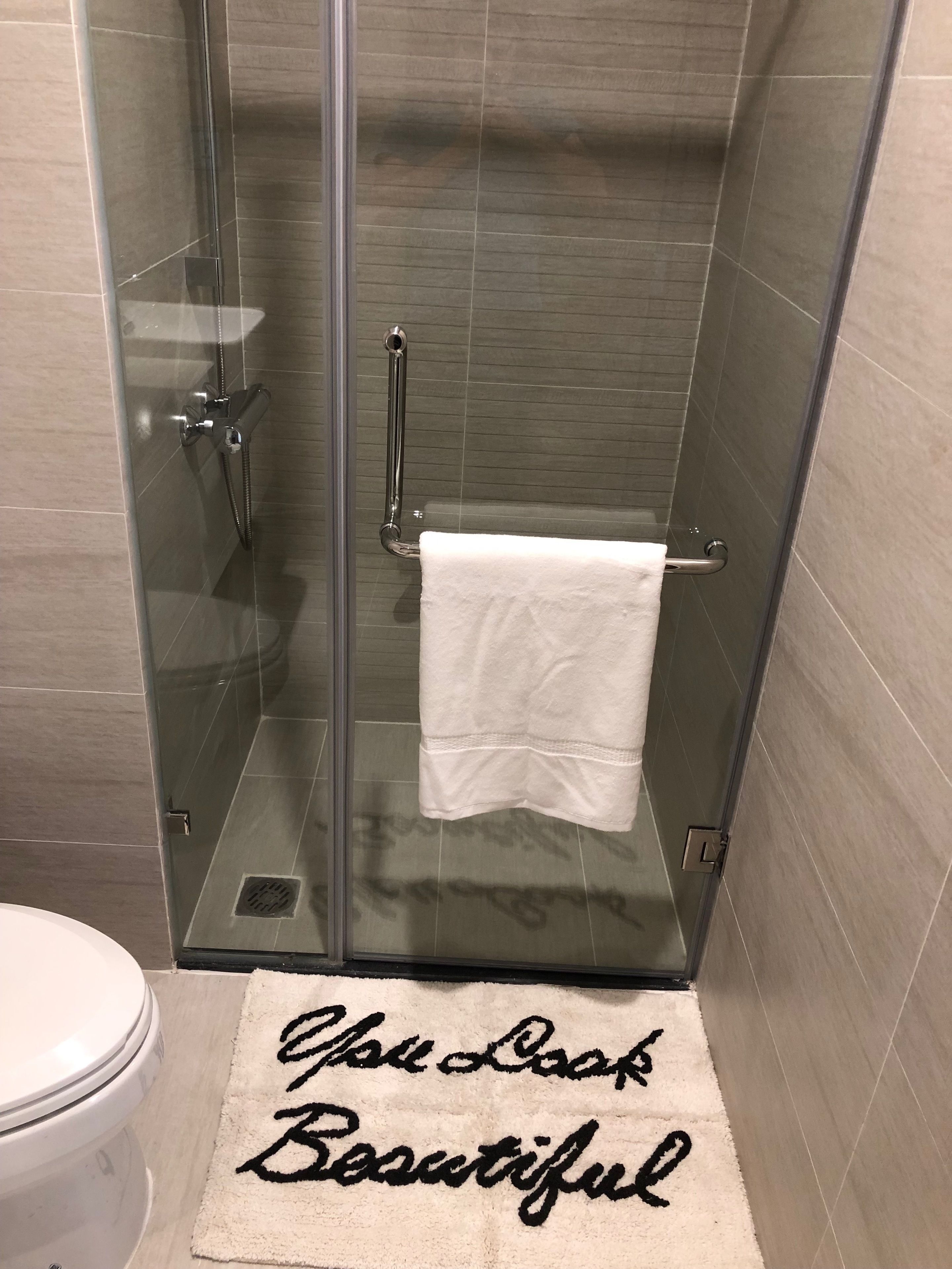En dusch med glasväggar, där en vit handduk hänger på en krok, en glasdörr och ett badrumsgolv med en matta som det står "Du lagar fantastisk mat" på.