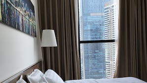 A hotel room with a large bed, a nightstand, a lamp, and a view of tall buildings through a window with curtains. - Luxury Condotel Vinhomes Skylake Keangnam (Hanoi)
