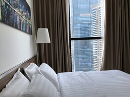 Habitación de hotel con una cama grande, una mesita de noche, una lámpara y vistas de edificios altos a través de una ventana con cortinas.
