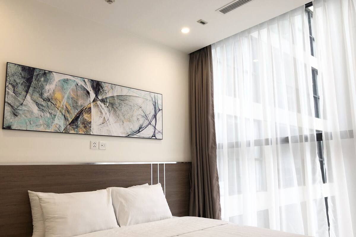 Une chambre d’hôtel avec un grand tableau abstrait au mur, un lit avec du linge de maison blanc, une tête de lit en bois et des rideaux légers à la fenêtre.
