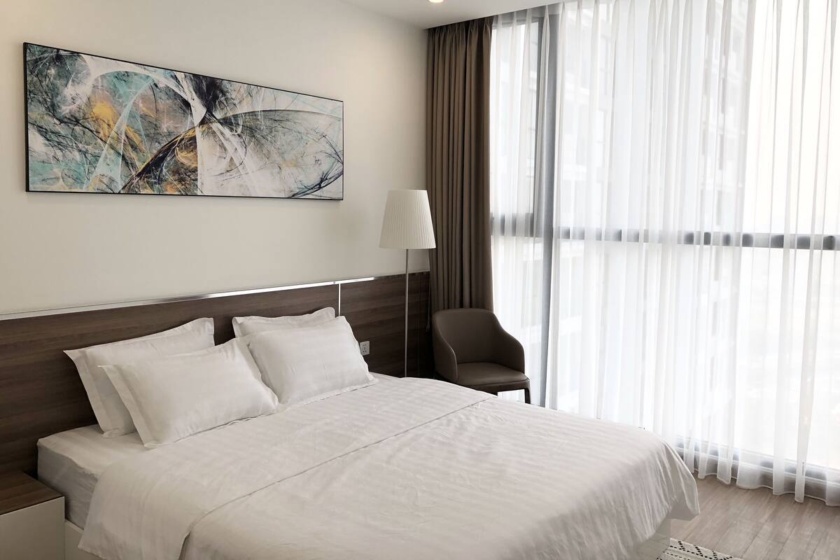 Une chambre d’hôtel avec un grand lit, une chaise, une lampe et une fenêtre avec des rideaux légers.