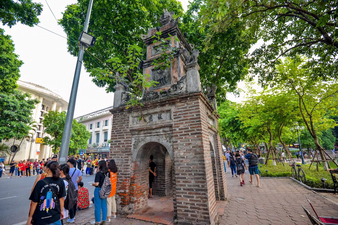 Monumento histórico de piedra con un arco, rodeado de árboles y una calle concurrida llena de gente.