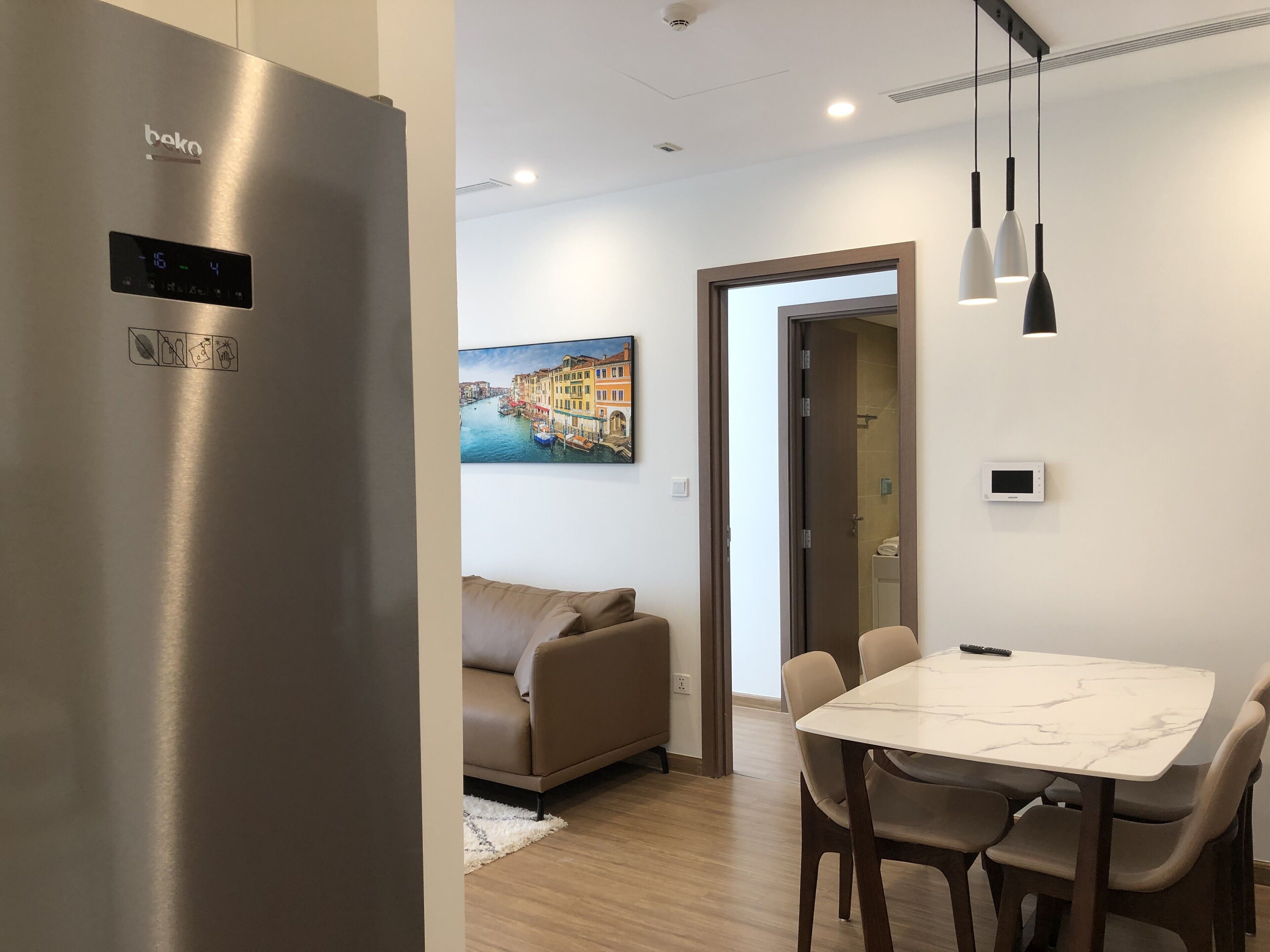 A modern kitchen with a stainless steel refrigerator, a dining area with a marble table and chairs, a beige sofa, and a painting of a cityscape on the wall.