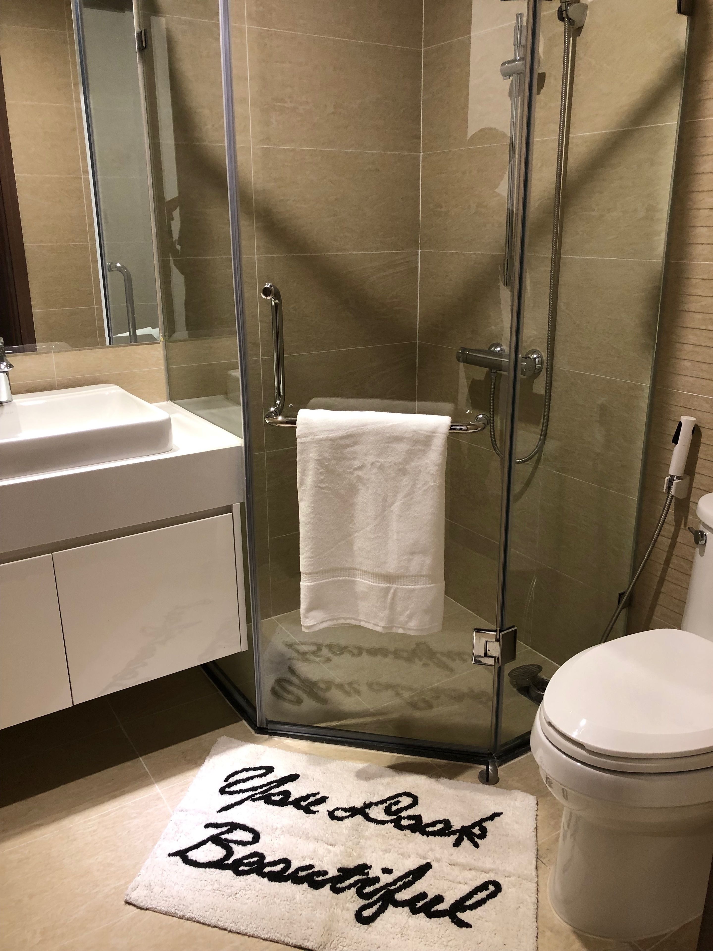 A bathroom with a glass-enclosed shower, a white sink, and a toilet. A towel hangs on the shower door, and a rug with the words "The Look Beautiful" is on the floor.