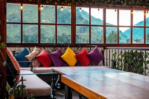 Une pièce avec vue sur des collines verdoyantes, grâce à de grandes fenêtres, un banc agrémenté de coussins colorés et une table.