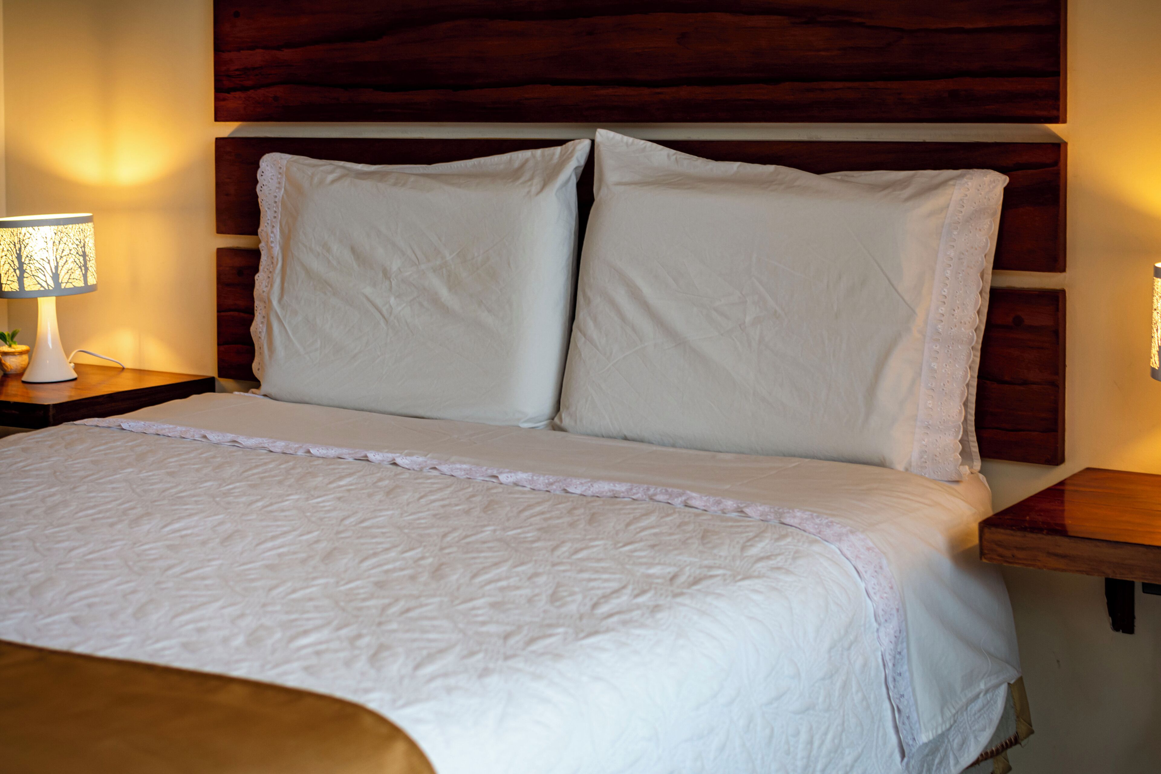 Una cama bien hecha con cabecera de madera, una mesita de noche con lámpara y una estantería en la pared.