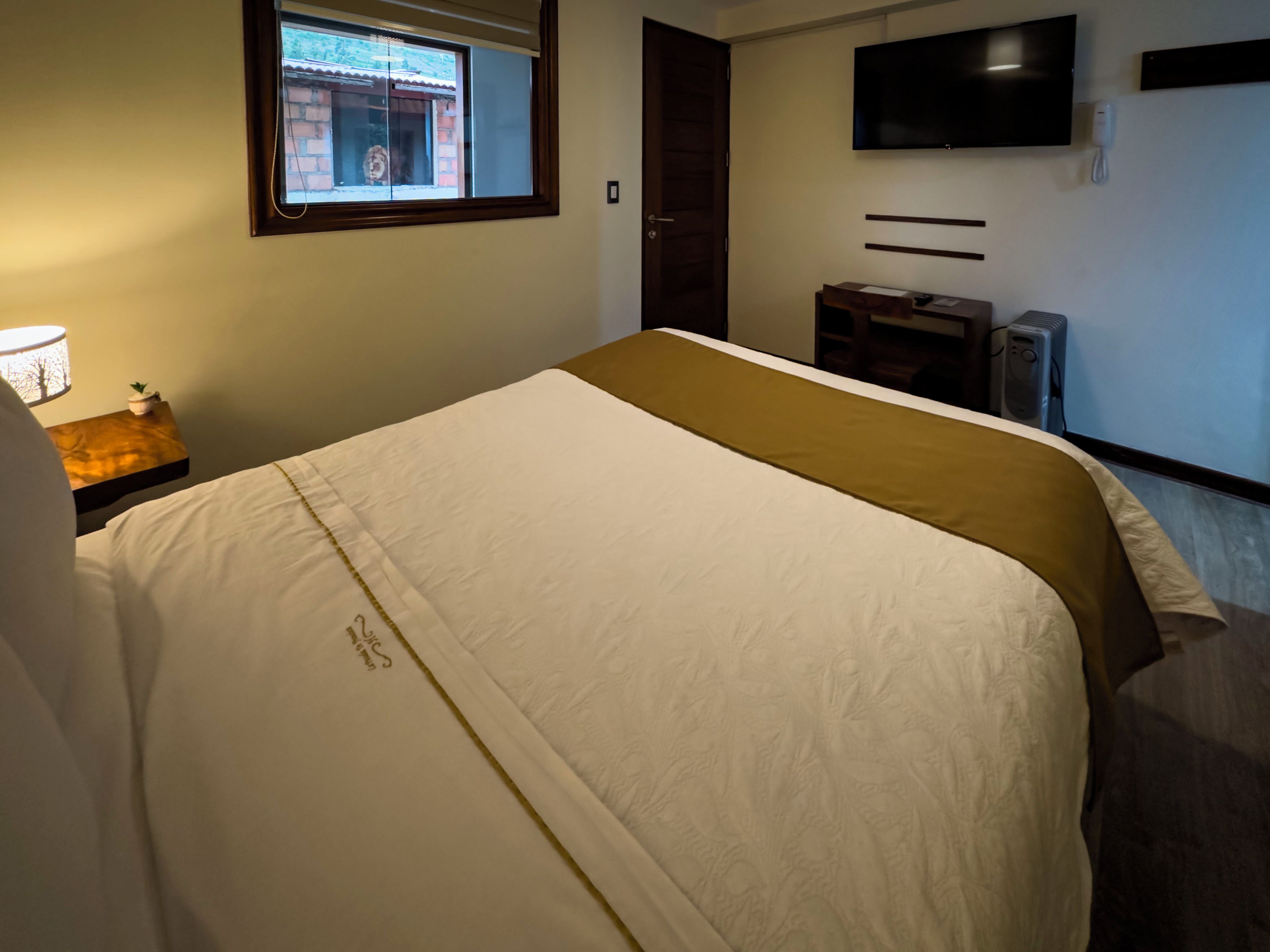 A hotel room with a bed, a bedside table with a lamp, a TV mounted on the wall, and a window with a view of a building.