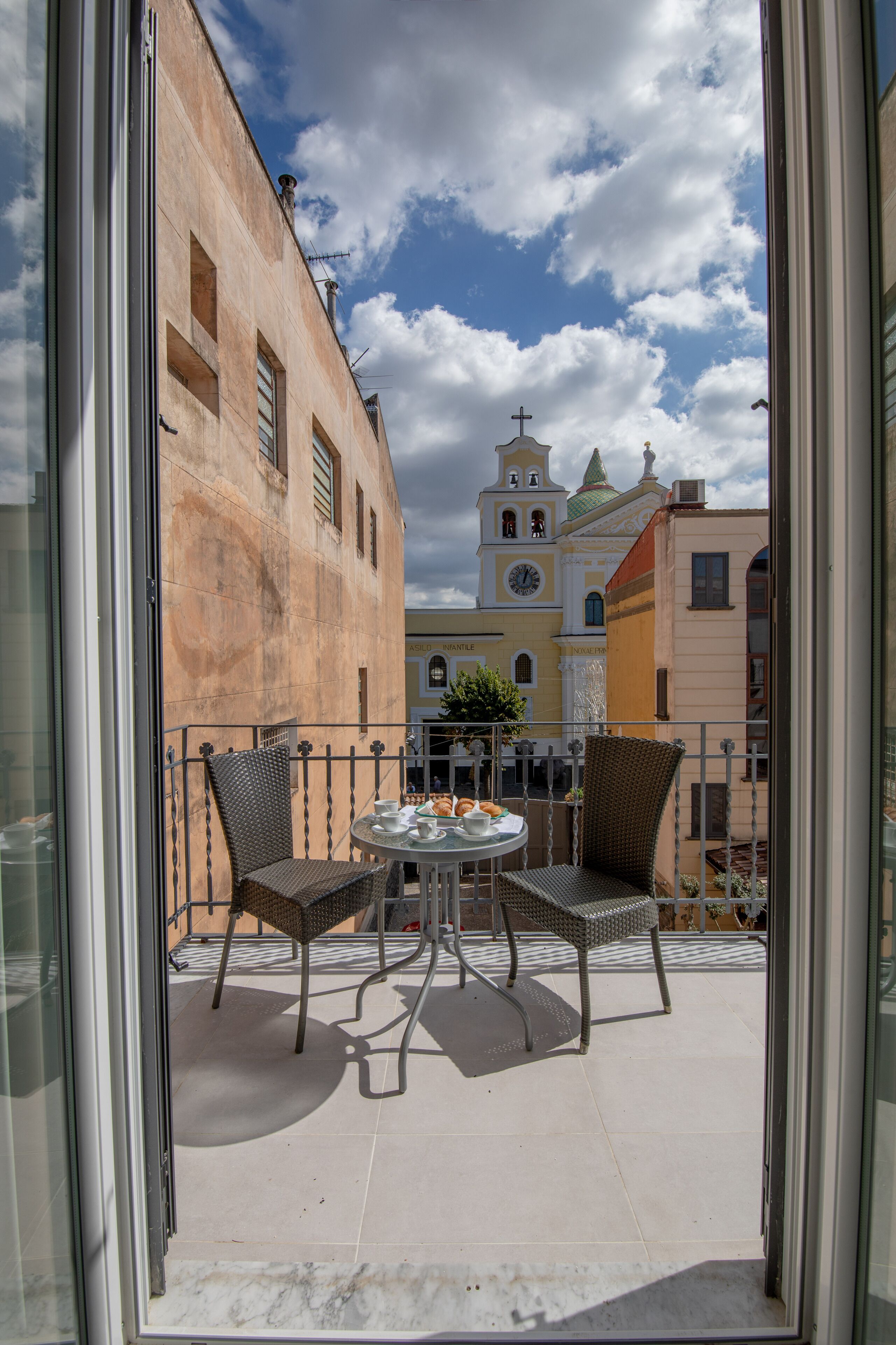 A balcony with two wicker chairs and a small table, overlooking a street with buildings and a church tower.