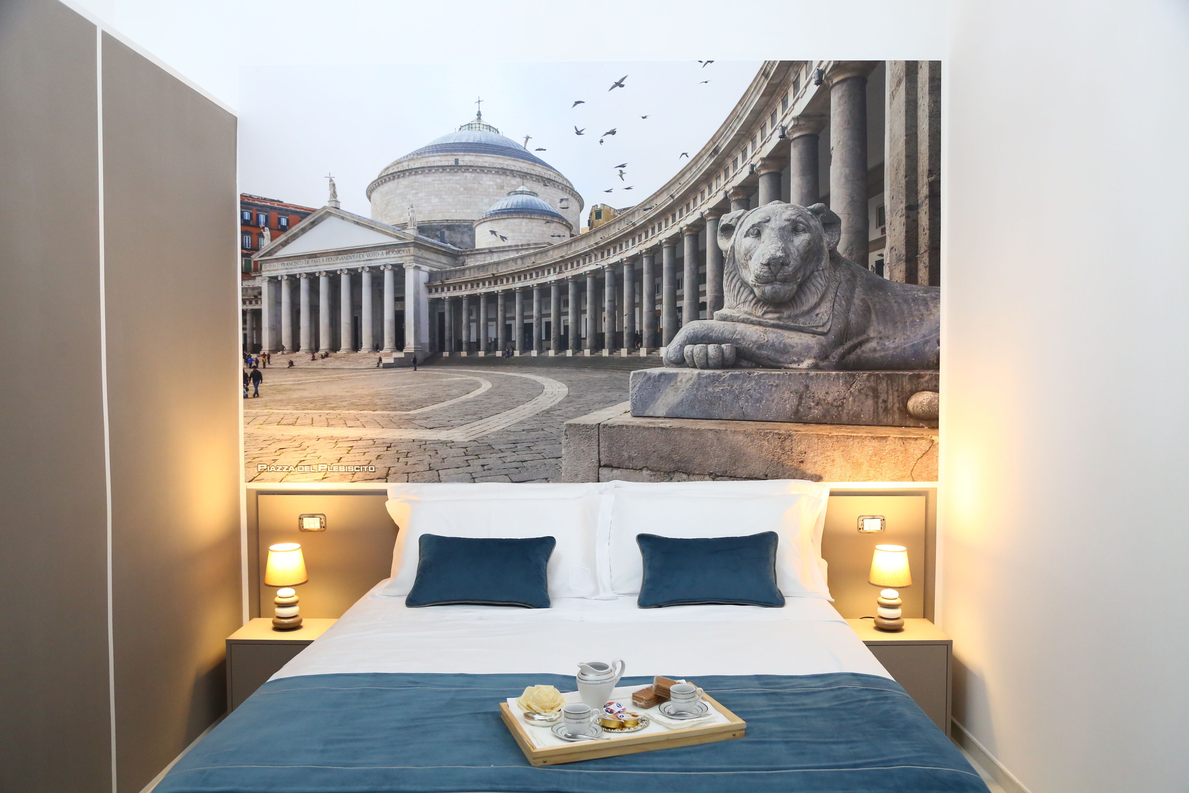 A hotel room with a bed, two pillows, a tray with a teapot and cups, and a view of a historic building with columns and a lion statue.