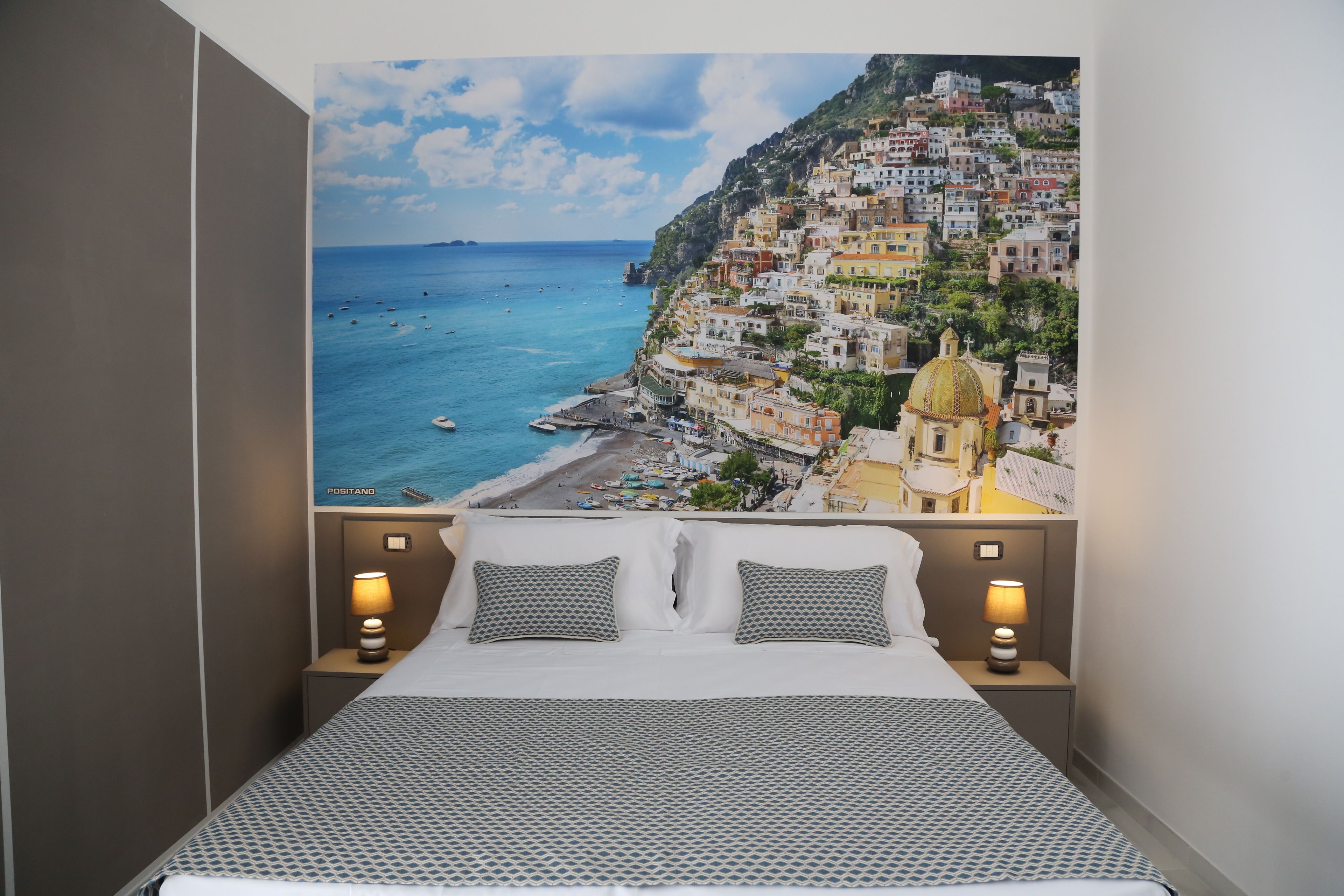 Un dormitorio con una cama, dos lámparas de noche y un amplio mural en la pared de un pueblo costero.