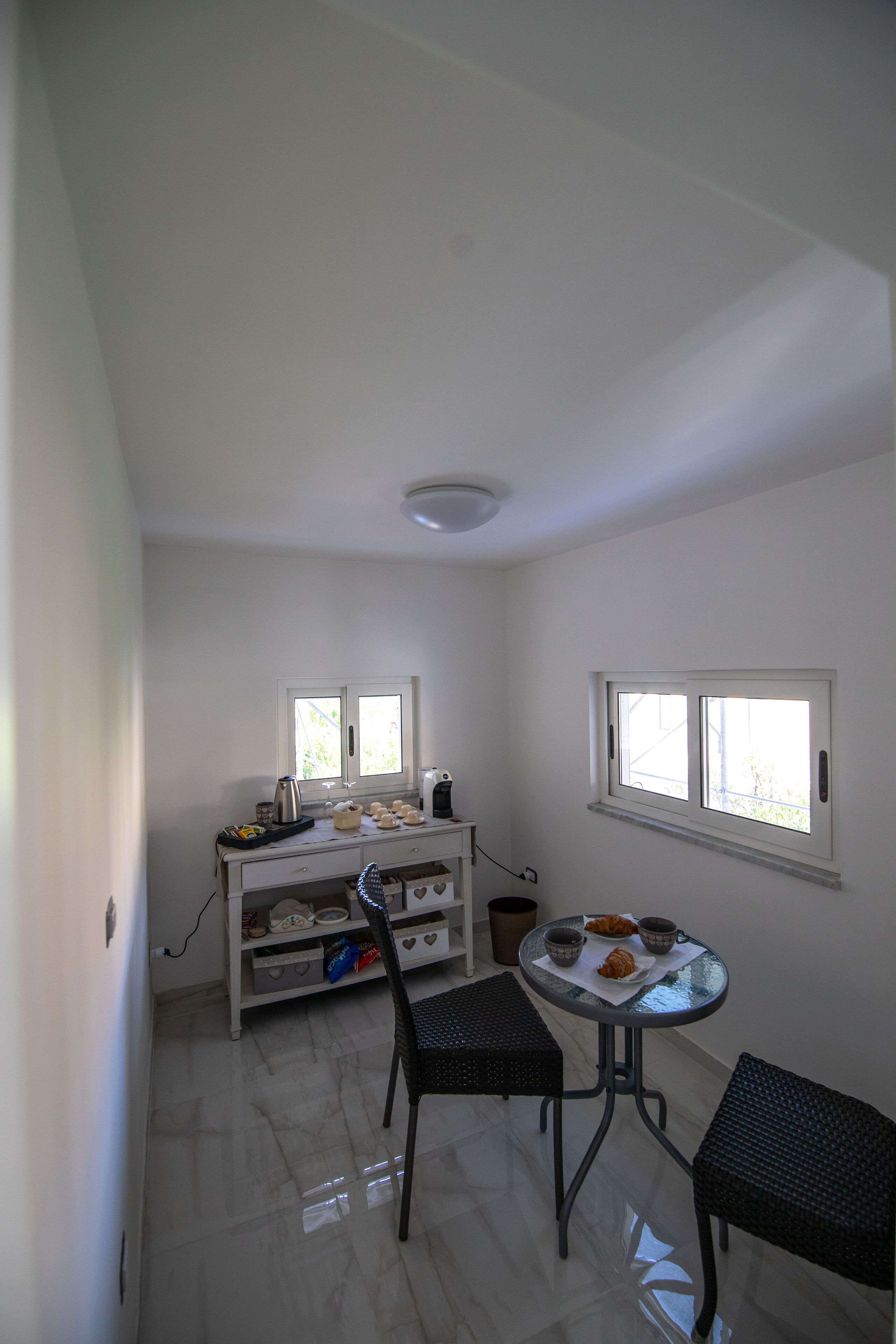 Una stanza piccola e pulita con un muro bianco, una finestra, uno scaffale bianco con diversi oggetti, un tavolo rotondo con sedie e un pavimento piastrellato.