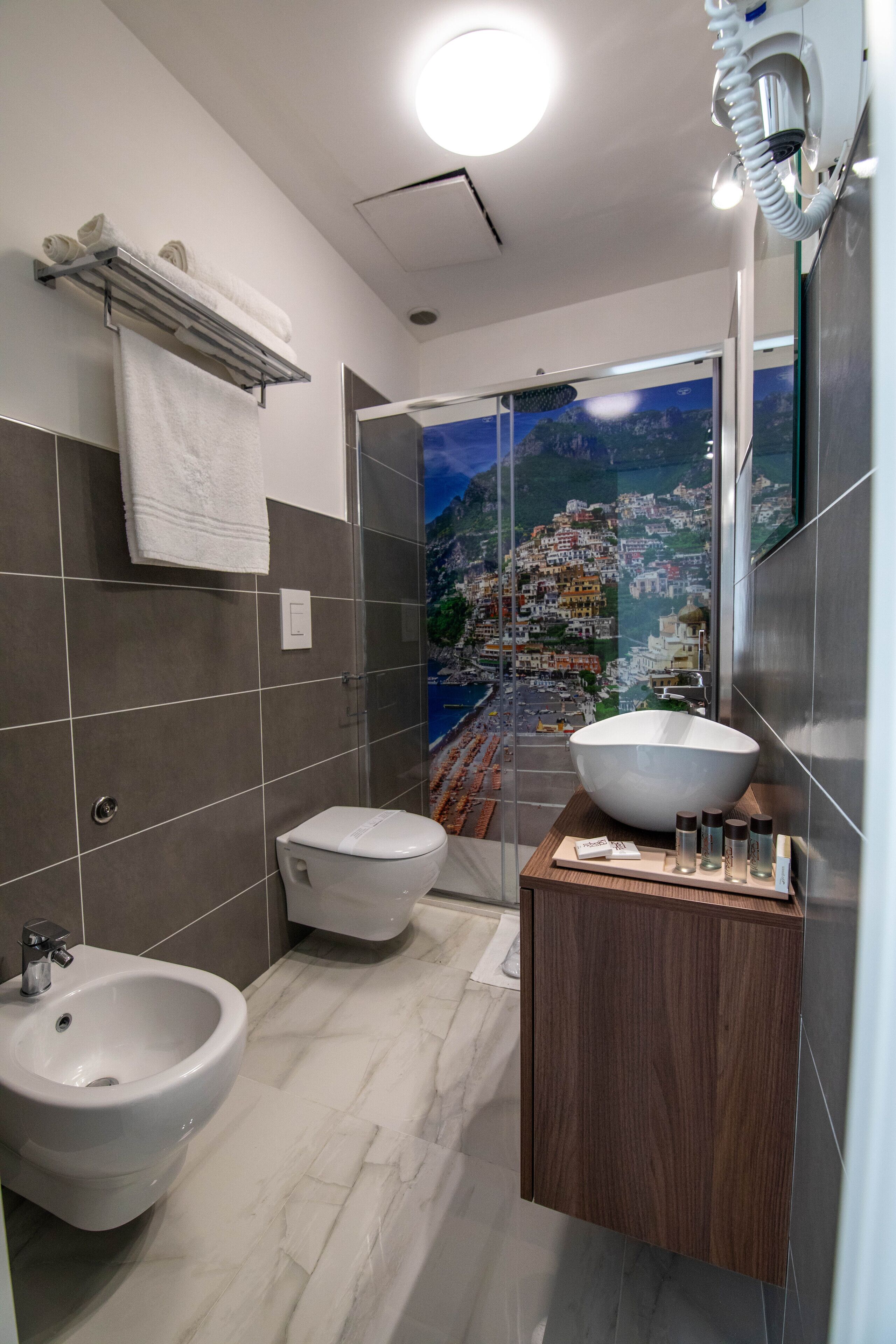 Une salle de bain moderne équipée d’un bidet, d’un grand lavabo et offrant une vue sur une ville côtière grâce à une porte vitrée.
