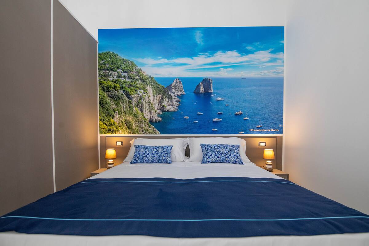 A hotel room with a bed featuring a large scenic view of a coastal landscape, including cliffs and boats.