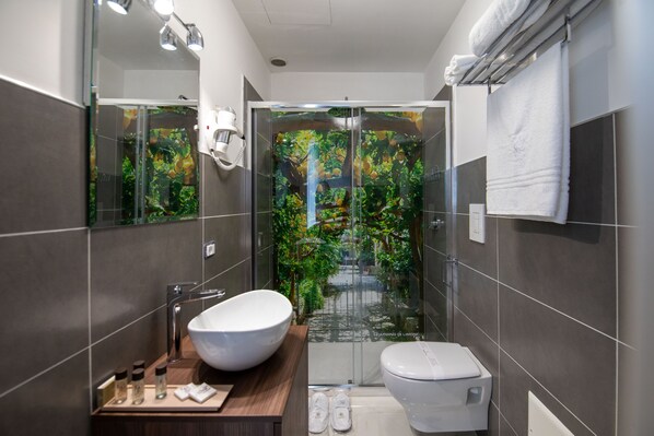 Een moderne badkamer met een groot wit wastafel, een toilet en een douche met glazen wanden met uitzicht op groen.