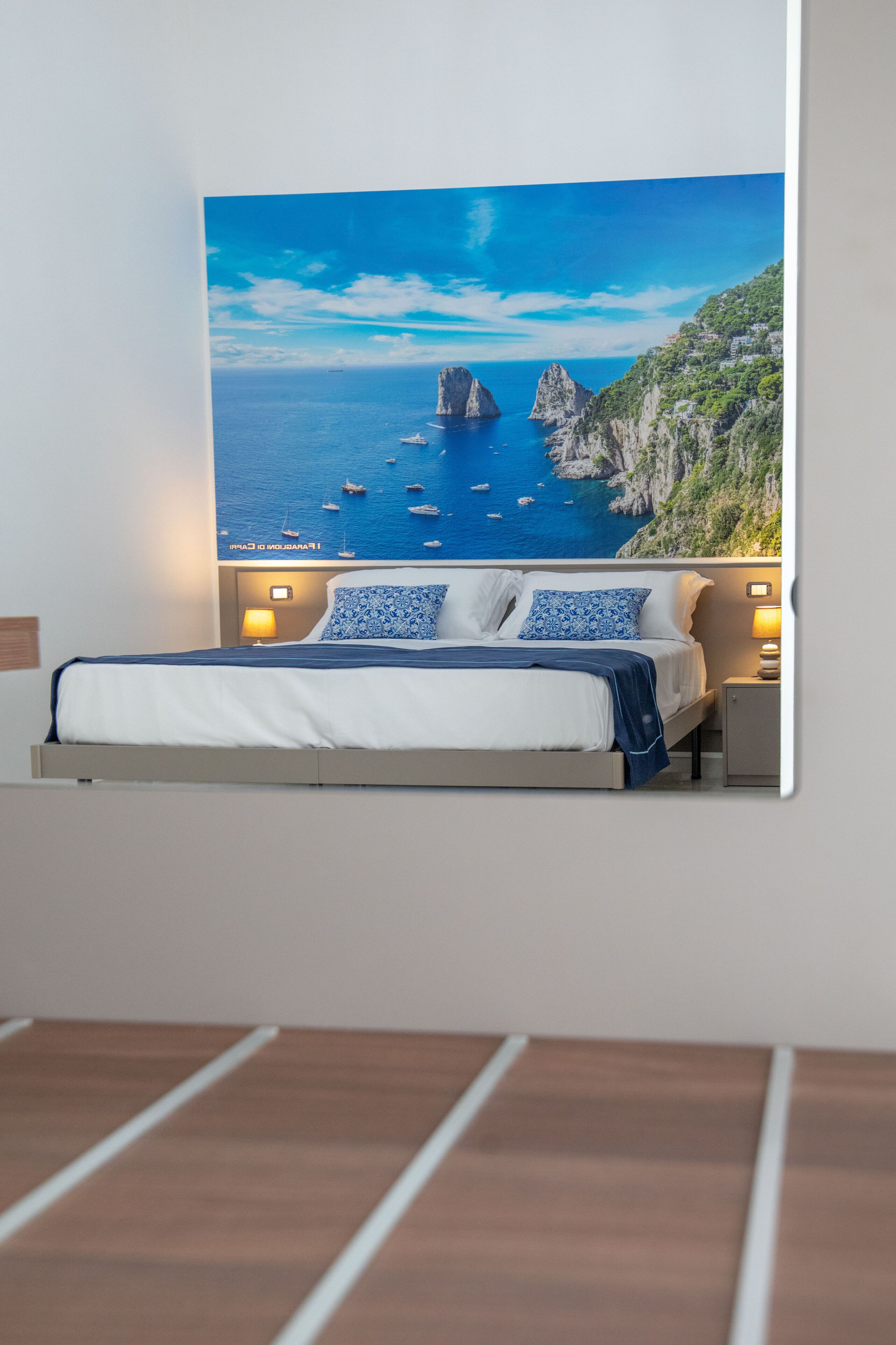 A bedroom with a bed, two bedside lamps, and a large painting of a coastal scene.