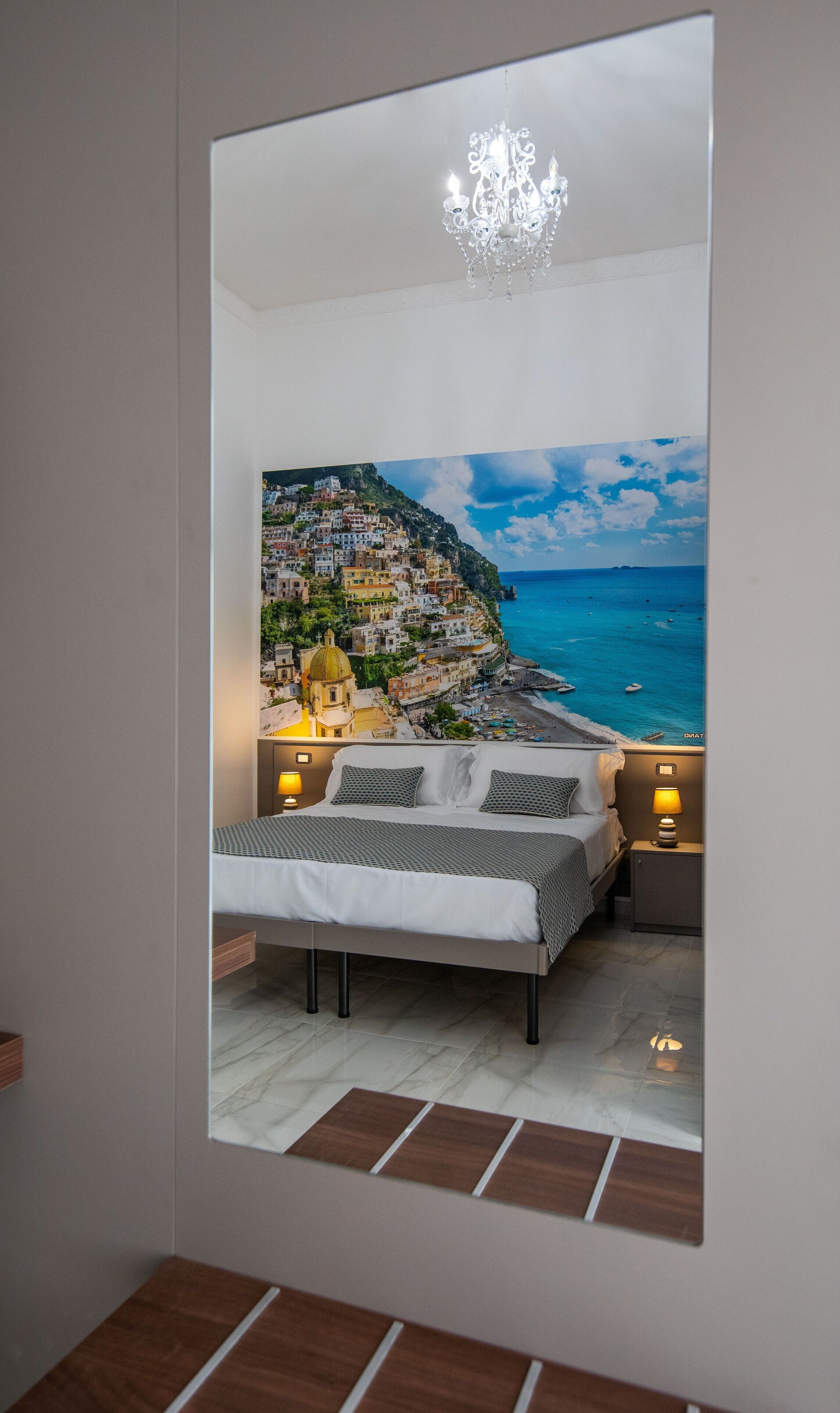 A bedroom with a bed, bedside lamps, a chandelier, and a large mirror reflecting a coastal town view.