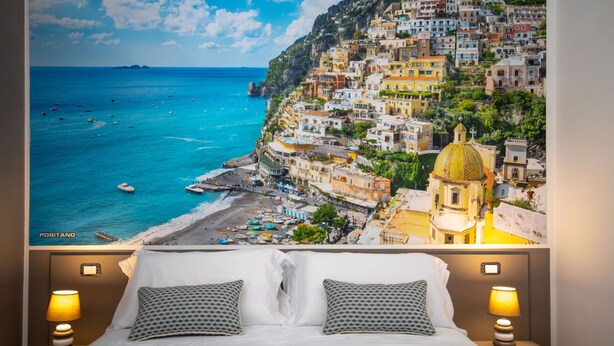 Habitación de hotel con cama, dos lámparas de noche y un cartel grande de un pueblo costero con barcos y edificios coloridos.