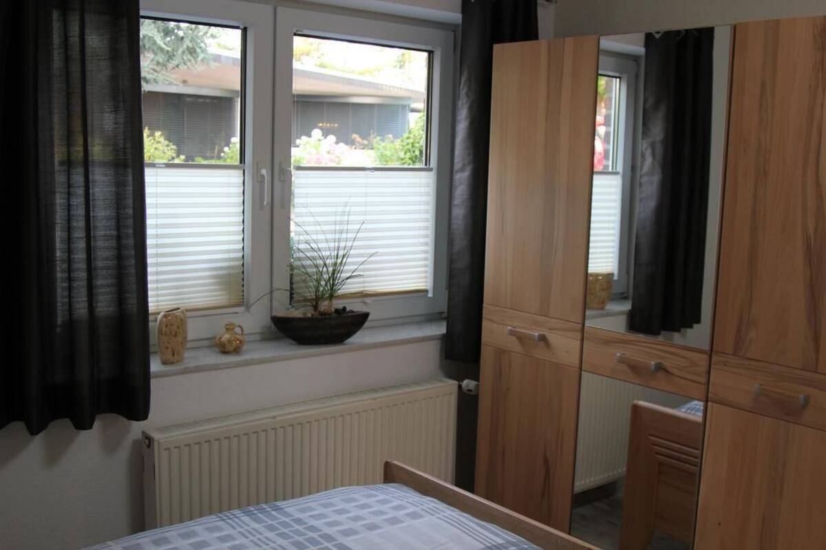 A bedroom with a bed, a window with blinds, a radiator, a wooden wardrobe, and a potted plant on the windowsill.