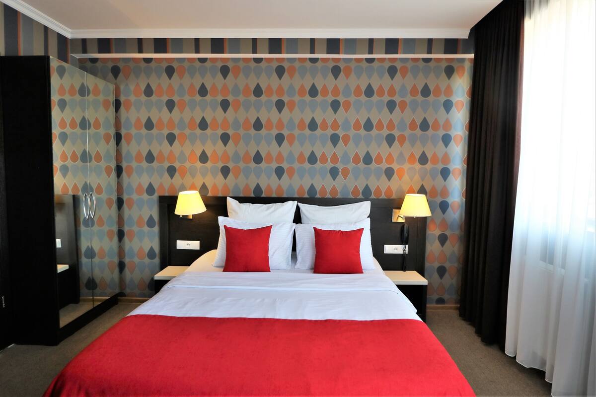 Habitación de hotel con una cama grande, dos mesitas de noche con lámparas, un espejo y un papel tapiz estampado.
