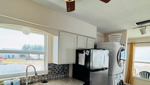 A compact kitchen with white cabinets, a stainless steel refrigerator, a washing machine, a ceiling fan, and a window providing a view of the outdoors. - 763 North Shore Upstairs 3 Bedroom Apts by RedAwning (Anna Maria)
