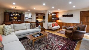 A spacious living room with a large sofa, a brown leather armchair, and a coffee table. There is a kitchen area with wooden cabinets and a hanging light fixture. - Texas Townhome Condos - 3 Minutes Walk to Gondola by RedAwning (Vail)