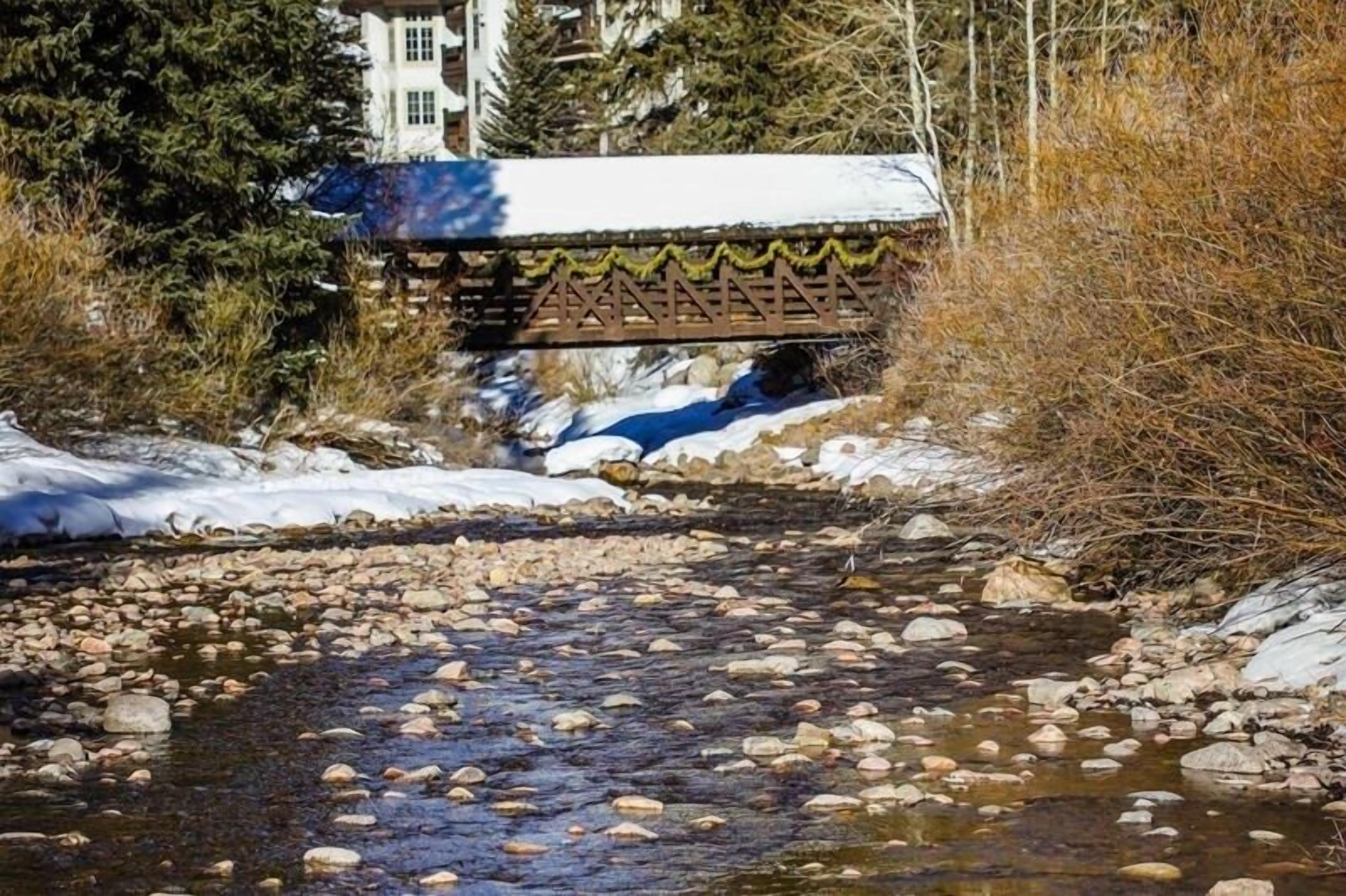 A bridge over a stream with snow on the ground and trees in the background.