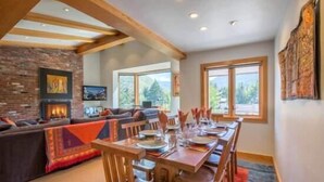 A dining area with a long wooden table set for six, a brick fireplace, and a large window offering a view of trees. - Mill Creek Court Condos Just 100 yards from Gondola by RedAwning (Vail)