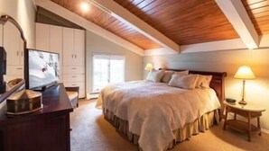 Un dormitorio con techo de madera, una cama con cubrecama blanco, una mesita de noche con lámpara y un aparador con espejo.