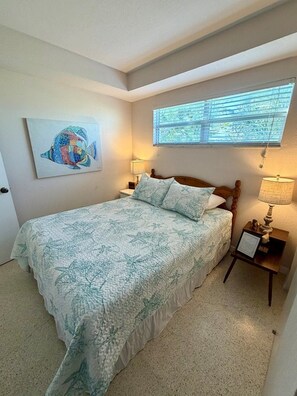 A bedroom with a bed, a window, a painting, and a nightstand with a lamp.