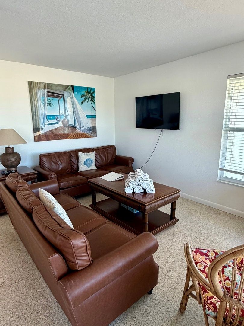 A living room with a brown leather sofa, a wooden coffee table, a flat-screen TV mounted on the wall, and a window with blinds.