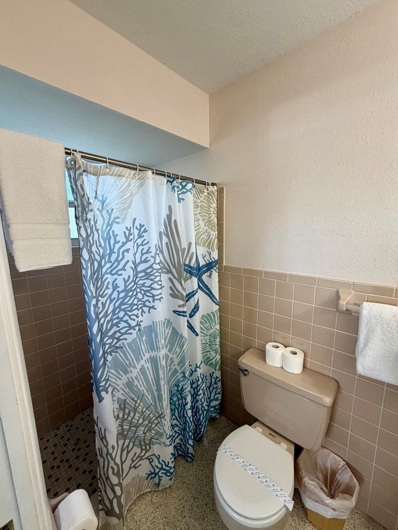Un baño con cortina de ducha estampada con motivos de coral, un inodoro con dos rollos de papel higiénico y un cubo de basura.