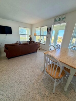 A living room with a brown sofa, a wooden dining table with white chairs, a TV mounted on the wall, and a sign that says 'BEACH' on the door.