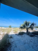 A view from under a structure with a palm tree and a distant tree, sandy ground, and a clear blue sky.