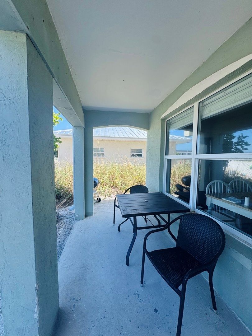 A covered patio with a table and chairs, a window with a view of a building, and a concrete floor.