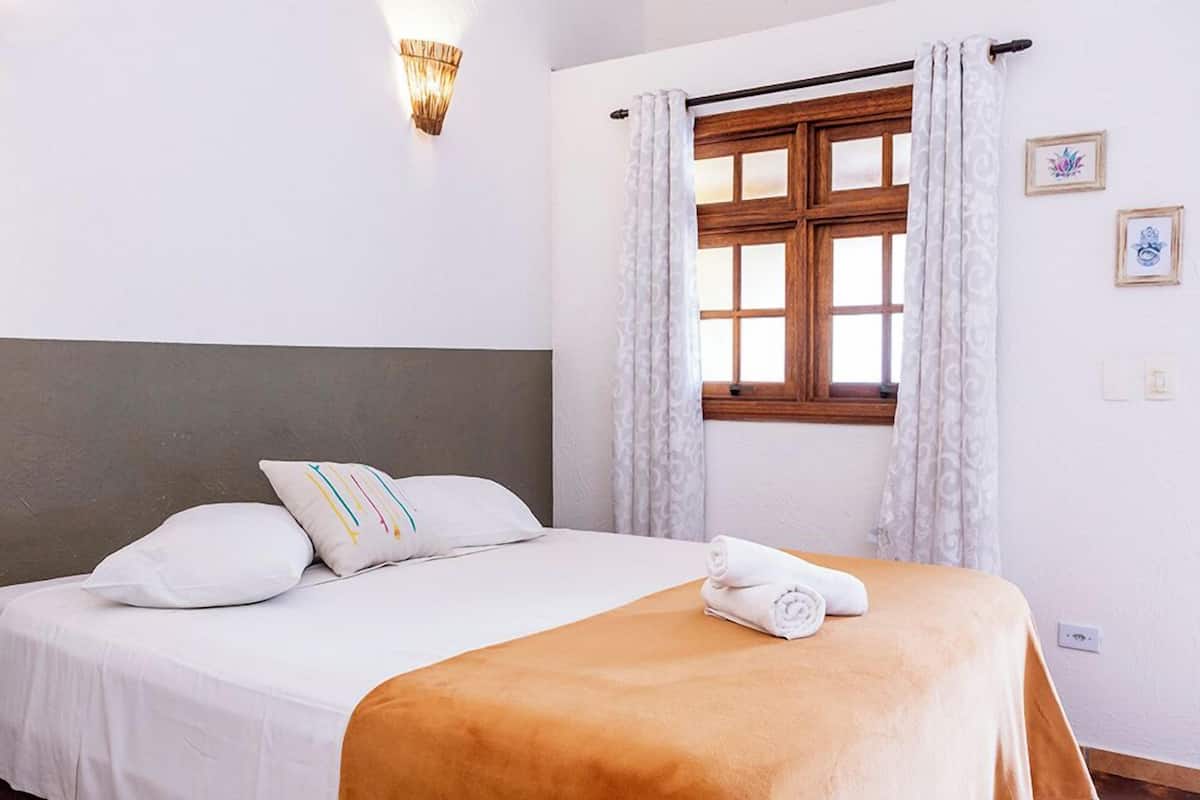 A neatly made bed with a white and brown bedspread, a pillow with colorful stripes, and a rolled towel on the bed. There is a window with wooden frames and white curtains, a wall-mounted light fixture, and two framed pictures on the wall.