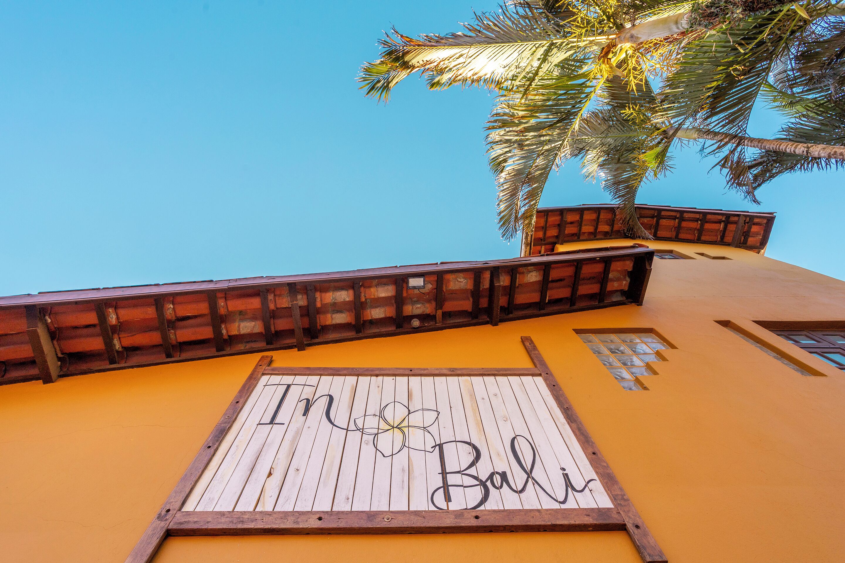 Uma placa de madeira com a inscrição 'In Bali', presa a um edifício com guarda-corpo de madeira e uma palmeira ao fundo.