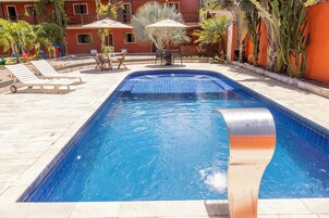Una piscina con sillones de descanso y sombrillas, rodeada de palmeras y un edificio con paredes rojas.