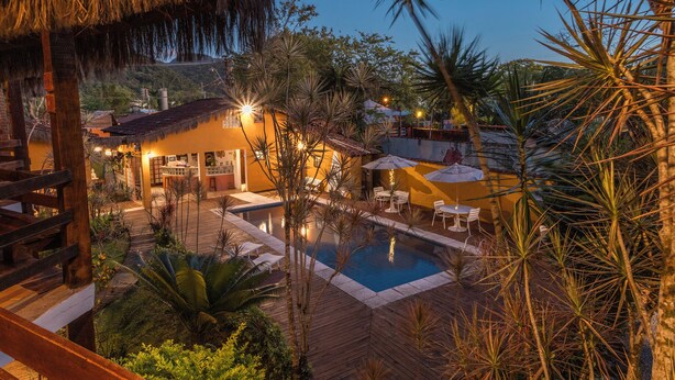 A pool area with a wooden deck, lounge chairs, and a thatched-roof building. - Pousada In Bali (São Sebastião)