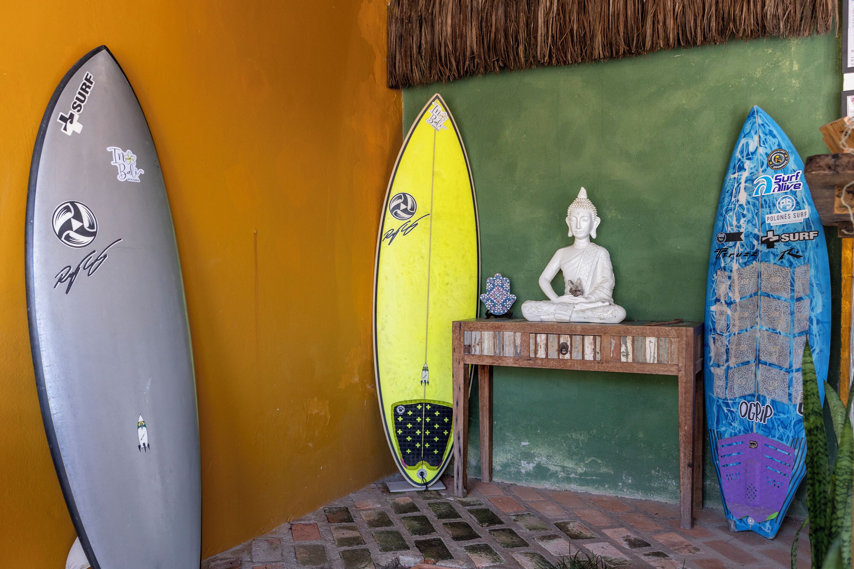 Tres tablas de surf apoyadas contra una pared, con una estatua de Buda y una mesa de madera al frente.
