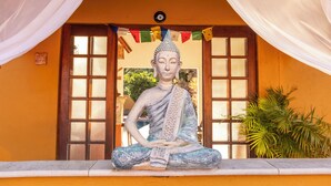 A statue of a seated figure in a meditative pose, set against a backdrop of a building with large windows and a curtain. - Pousada In Bali (São Sebastião)