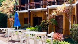 A two-story building with a pool area featuring white plastic chairs and a blue umbrella. - Pousada In Bali (São Sebastião)