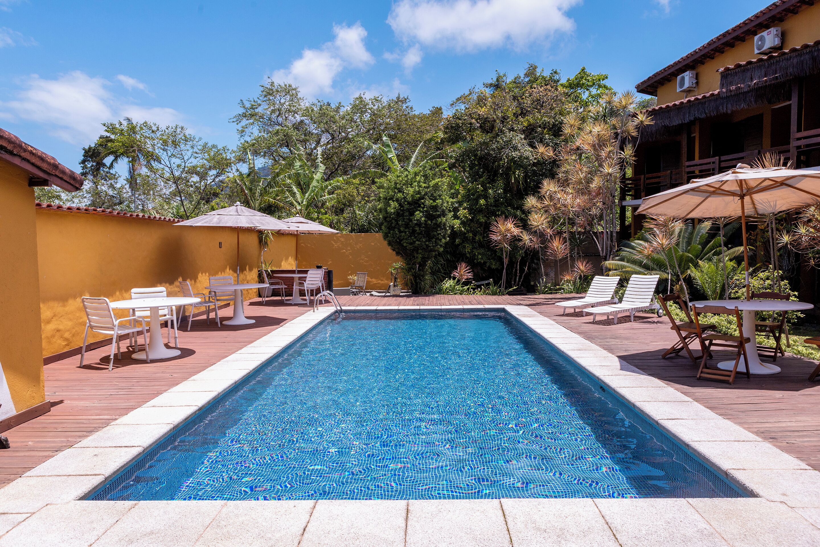 A rectangular swimming pool with tiled edges, surrounded by a wooden deck. There are white lounge chairs and tables with umbrellas, and a building with a thatched roof in the background.